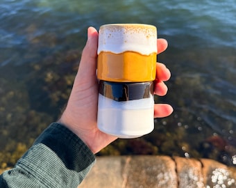May include: A set of two stacked ceramic cups. The top cup features a white rim with a speckled texture transitioning into a solid mustard yellow base. The bottom cup has a glossy dark brown upper section and a smooth white lower base.