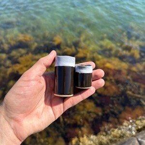 K&ouml;nnte beinhalten: Zwei zylindrische Keramikbecher mit einer gl&auml;nzend dunkelbraunen Oberfl&auml;che im unteren Bereich und einer strukturierten, mattwei&szlig;en Glasur am oberen Rand. Die Becher haben unterschiedliche H&ouml;hen, wobei einer deutlich gr&ouml;&szlig;er ist als der andere.