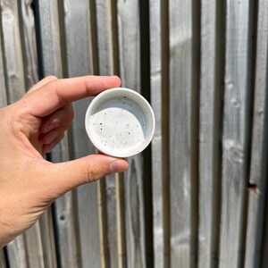 May include: A small, circular ceramic dish with a white glaze and a speckled pattern featuring tiny black dots scattered across the interior surface. The dish has a smooth, rounded rim and a shallow basin, presenting a clean and simple design.