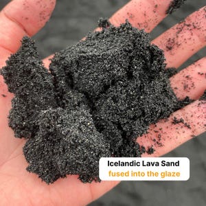May include: A coarse, dark black volcanic material with a granular, crystalline texture. A white rectangular label with rounded corners features the black text "Icelandic Lava Sand" and the orange text "fused into the glaze" printed on it.