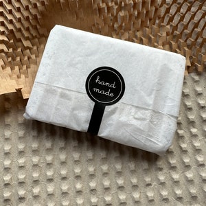 May include: A rectangular package wrapped in thin white paper, secured with a circular black sticker featuring the white text hand made. The sticker has a vertical tab extending downward, sealing the center of the paper wrapping.