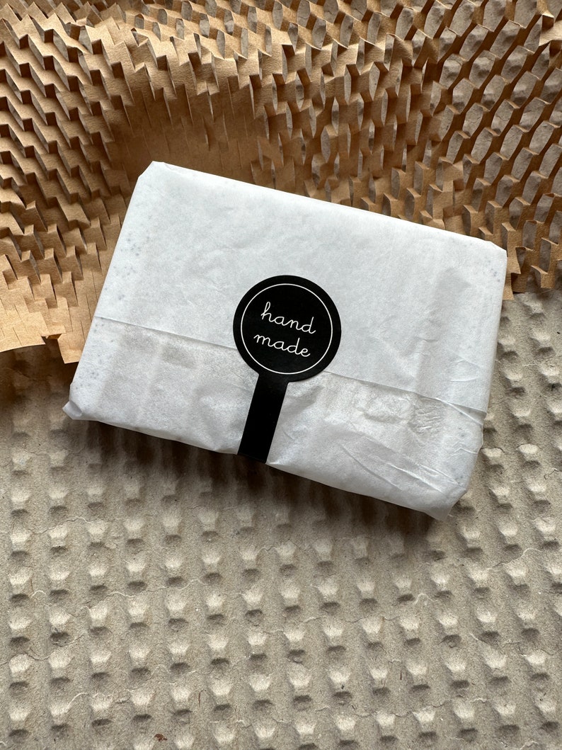 May include: A rectangular package wrapped in thin white paper, secured with a circular black sticker featuring the white text hand made. The sticker has a vertical tab extending downward, sealing the center of the paper wrapping.