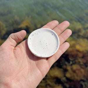 May include: A small, round, white ceramic dish featuring a speckled pattern with tiny black dots across its surface. The dish has a smooth, slightly concave center and a raised rim, designed as a compact tray for holding small items.