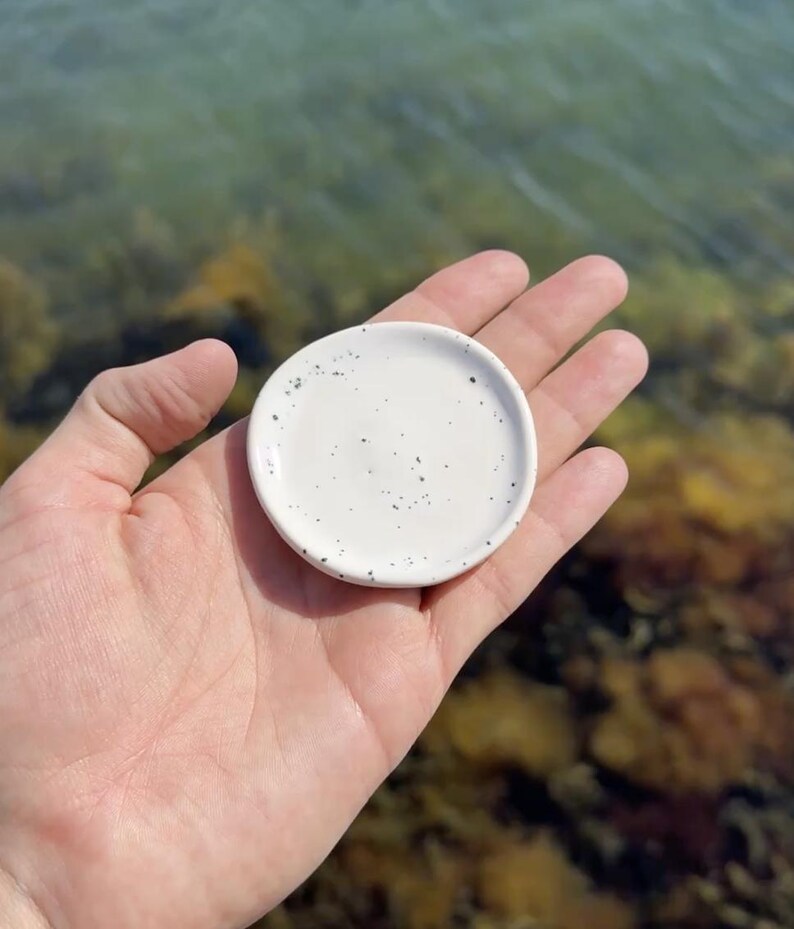 May include: A small, round, white ceramic dish featuring a speckled pattern with tiny black dots across its surface. The dish has a smooth, slightly concave center and a raised rim, designed as a compact tray for holding small items.
