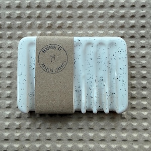 May include: A rectangular white ceramic soap dish with black speckles and vertical drainage grooves. A brown paper band wraps around the center, featuring a circular stamp that reads HANDMADE BY MAAKSEN CERAMICS with a central geometric logo.