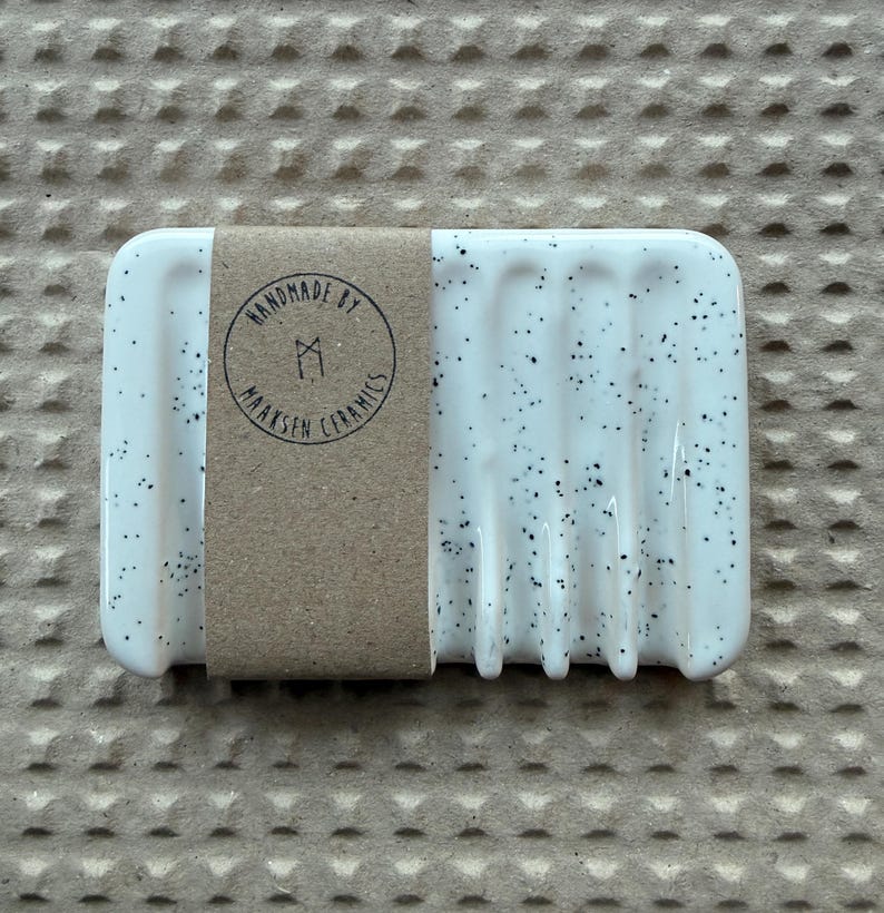 May include: A rectangular white ceramic soap dish with black speckles and vertical drainage grooves. A brown paper band wraps around the center, featuring a circular stamp that reads HANDMADE BY MAAKSEN CERAMICS with a central geometric logo.