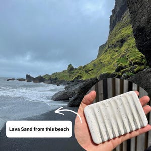 May include: A rectangular white soap dish featuring a speckled pattern of black dots and five parallel drainage grooves. The surface is smooth and the design includes a text overlay stating Lava Sand from this beach.