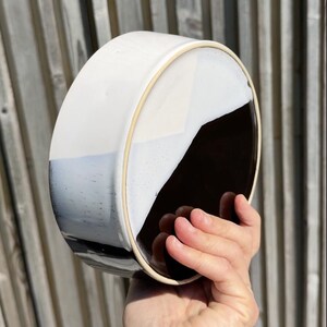 Könnte beinhalten: Zweifarbiges Keramikschale, oben weiß und unten dunkelbraun, mit hellem braunen Rand. Die Schale wird in einer Hand vor einem Holzzaun gehalten. Modernes und minimalistisches Design.