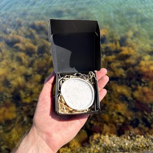 May include: A small, round, white ceramic dish with black speckles nestled inside a black cardboard gift box. The dish rests on a bed of light brown shredded paper filler, creating a contrast between the white ceramic and the dark box.