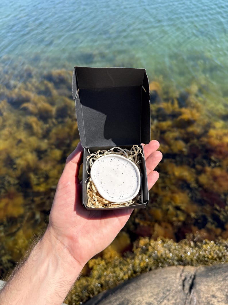 May include: A small, round, white ceramic dish with black speckles nestled inside a black cardboard gift box. The dish rests on a bed of light brown shredded paper filler, creating a contrast between the white ceramic and the dark box.
