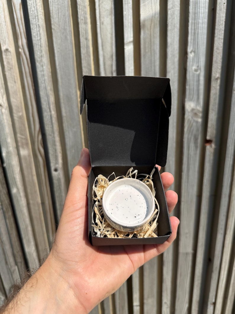 May include: A small, round, white ceramic bowl with black speckles nestled inside a black cardboard gift box. The bowl is surrounded by light-colored wood shavings used as protective packaging material within the open container.