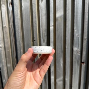 May include: A small, cylindrical ceramic dish featuring a white base with scattered black speckles throughout the exterior surface. The object has a smooth, flat rim and a shallow interior, showcasing a simple, functional design for holding small items.
