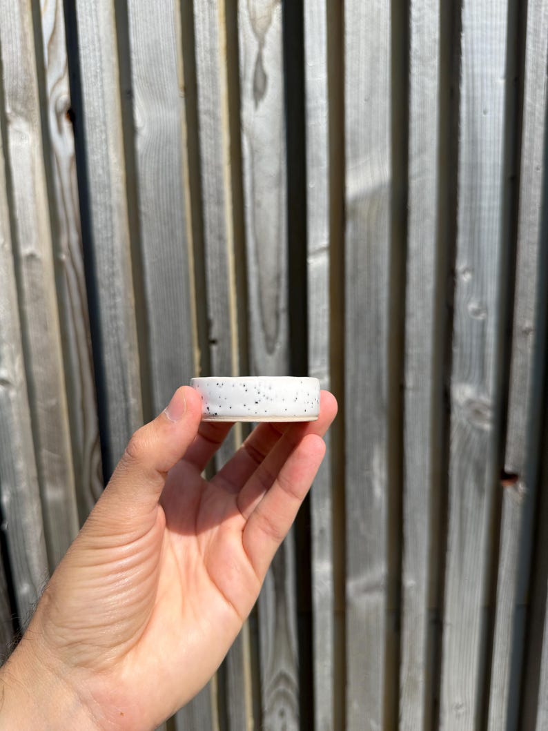 May include: A small, cylindrical ceramic dish featuring a white base with scattered black speckles throughout the exterior surface. The object has a smooth, flat rim and a shallow interior, showcasing a simple, functional design for holding small items.