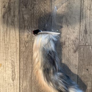 Puede incluir: Una cola de piel sintética en tonos blanco, gris y melocotón. La cola está sujeta a un clip negro y rojo, y está diseñada para ser usada como accesorio. La cola es esponjosa y tiene un efecto degradado.
