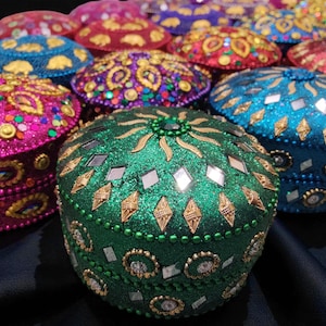 May include: A selection of colourful, round boxes, each heavily decorated with glitter, mirrored elements, and gold-coloured embellishments. The boxes are in shades of green, pink, blue, red, and purple. These are likely decorative or storage containers.