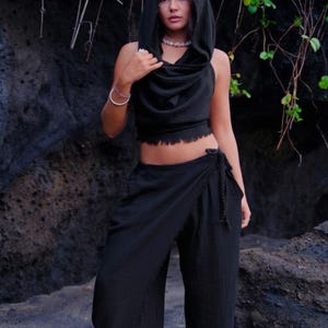 May include: Black hooded crop top and matching wide-leg pants. The top has a cowl neck and a raw hem. The pants are a wrap style with a tie at the waist. The outfit is made of a textured fabric.
