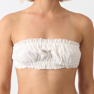 May include: A white, strapless tube top made of a textured fabric. The top is gathered at the top and bottom, creating a ruffled effect. The edges are unfinished, giving it a casual look. The top is worn by a person.