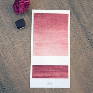 May include: A watercolor paint swatch card with a gradient of pink and red shades. The card is labeled "DUSK" and shows a darker red swatch below the gradient.