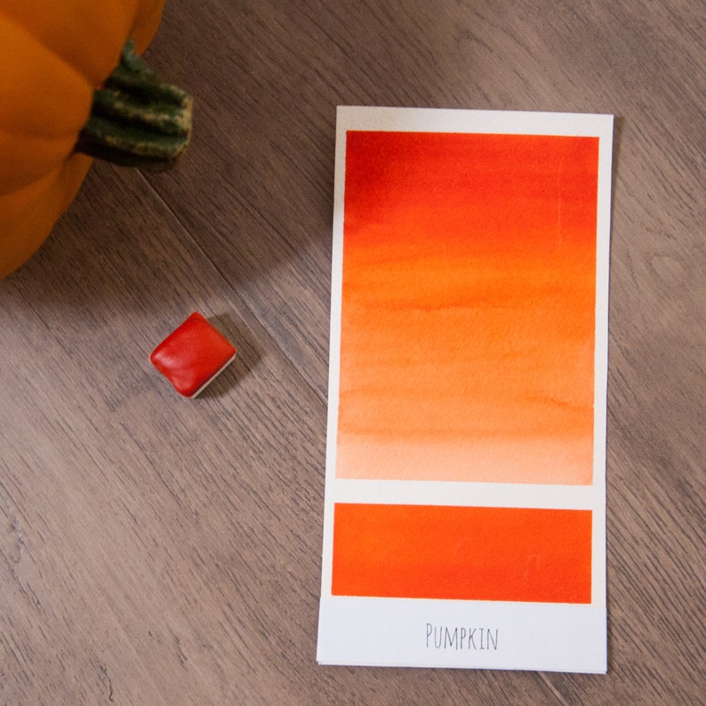 May include: A watercolor paint swatch card with a gradient of orange and red shades. The card is labeled "PUMPKIN" and shows a small square of solid orange paint.
