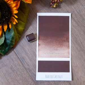 May include: A watercolor paint swatch in the color Roasted Chestnut. The swatch shows a gradient from a dark brown to a light brown. The paint is in a small, square pan.