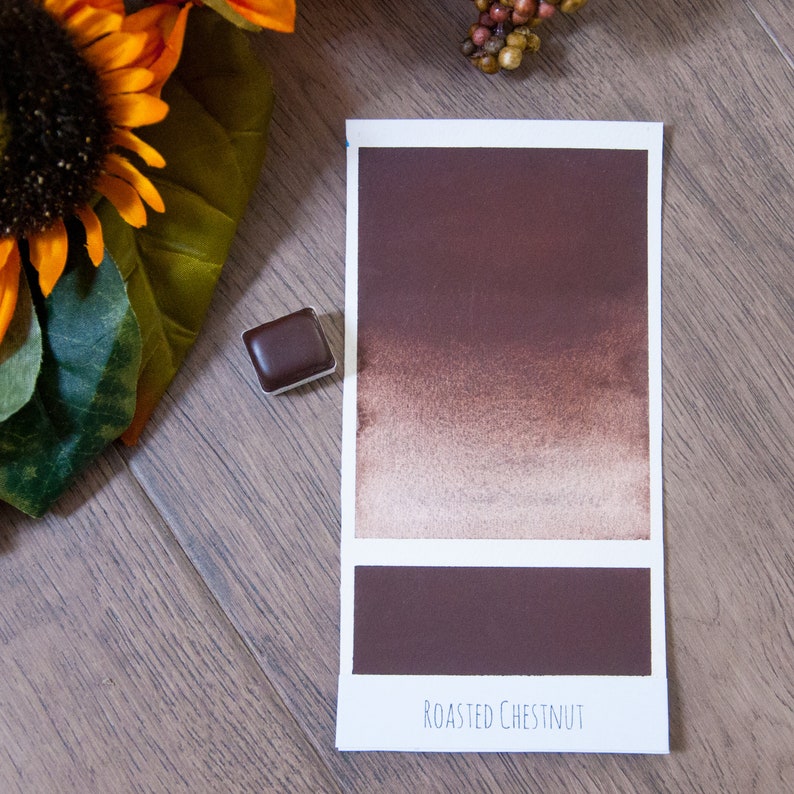 May include: A watercolor paint swatch in the color Roasted Chestnut. The swatch shows a gradient from a dark brown to a light brown. The paint is in a small, square pan.