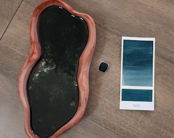 Può includere: Un vassoio di legno con un interno verde scuro riempito di resina e un bordo marrone chiaro. Sono presenti anche un piccolo oggetto nero quadrato e una scheda campione di acquerello con la parola "Thames". La scheda mostra una sfumatura di tonalità blu-verdi.