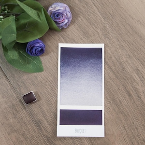 May include: A watercolor paint pan with a gradient of purple shades, from dark to light, with the word "BOUQUET" printed below. The pan is on a wooden surface with a small pan of brown paint and purple flowers.