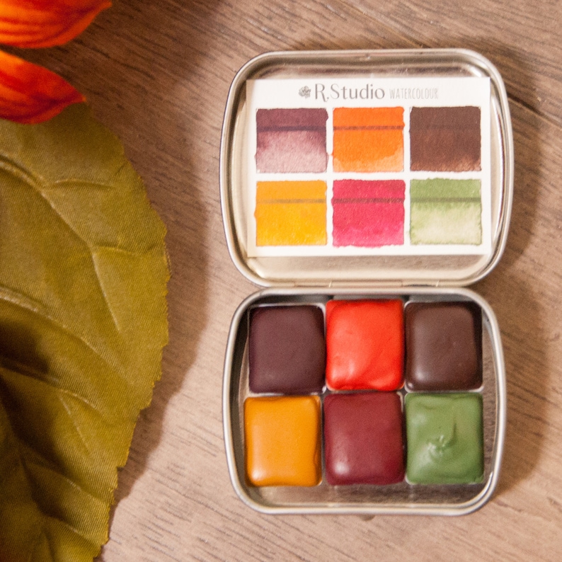 May include: A metal tin of R.Studio watercolor paints. The tin contains six watercolor pans in shades of brown, orange, red, and green. The tin is open and the pans are visible.