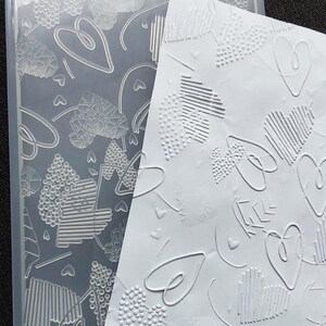 Könnte beinhalten: Ein transparenter Prägeordner und ein weißes geprägtes Papier, beide mit Herzmustern. Die Herzen weisen verschiedene Texturen auf, darunter gepunktete, linierte und umrissene Designs, die einen taktilen und optisch ansprechenden Effekt erzeugen.