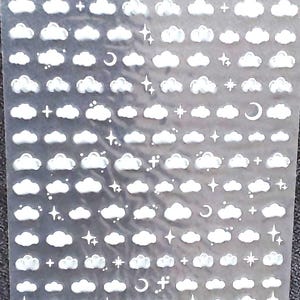 Könnte beinhalten: Eine Folie mit weißen Wolken-, Stern- und Halbmond-Nagelaufklebern. Die Folie ist transparent und hat den Text "Aha! 025" aufgedruckt.