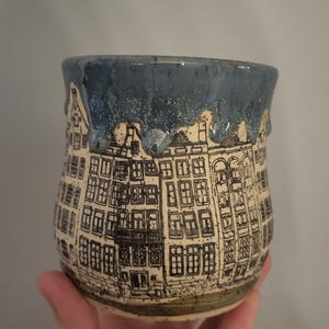 May include: A ceramic mug with a hand-drawn illustration of buildings. The mug features a blue rim and a beige body with black line art depicting windows and architectural details. The mug is held by a hand.
