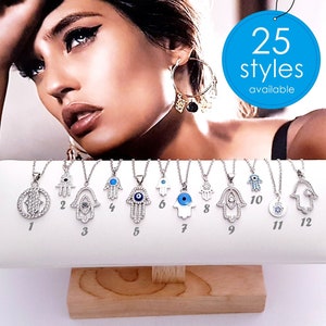May include: Twelve silver Hamsa hand necklaces with blue evil eye charms and crystal accents displayed on a white stand. The necklaces are numbered 1 through 12. A sign above the necklaces reads "25 styles available".