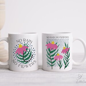 Może przedstawiać: Dwa białe ceramiczne kubki z kwiatowym wzorem i tekstem "No rain no flowers". Jeden kubek ma jeden kwiat, a drugi trzy kwiaty.