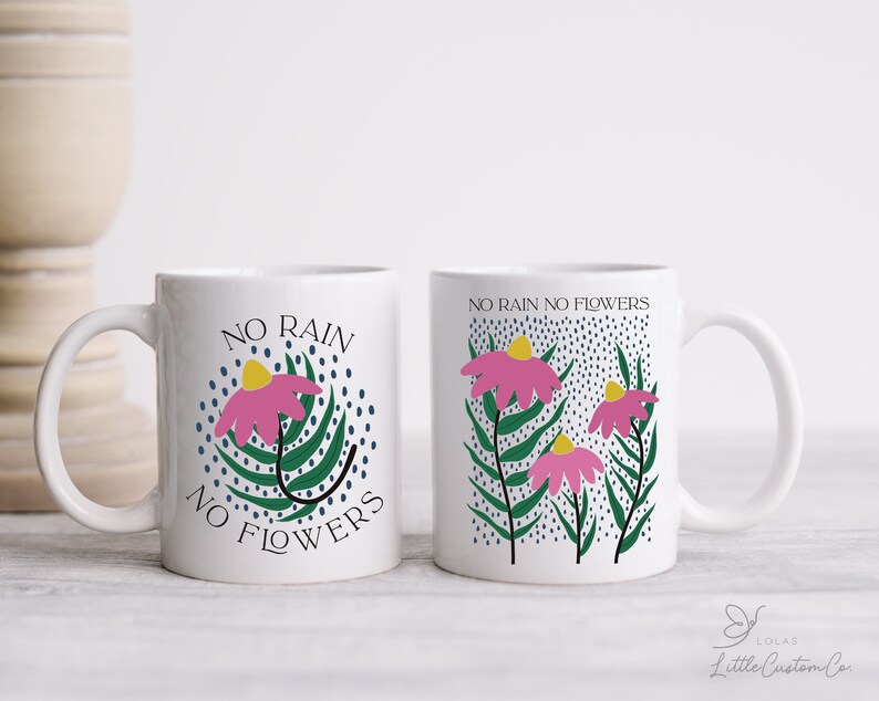 Może przedstawiać: Dwa białe ceramiczne kubki z kwiatowym wzorem i tekstem "No rain no flowers". Jeden kubek ma jeden kwiat, a drugi trzy kwiaty.