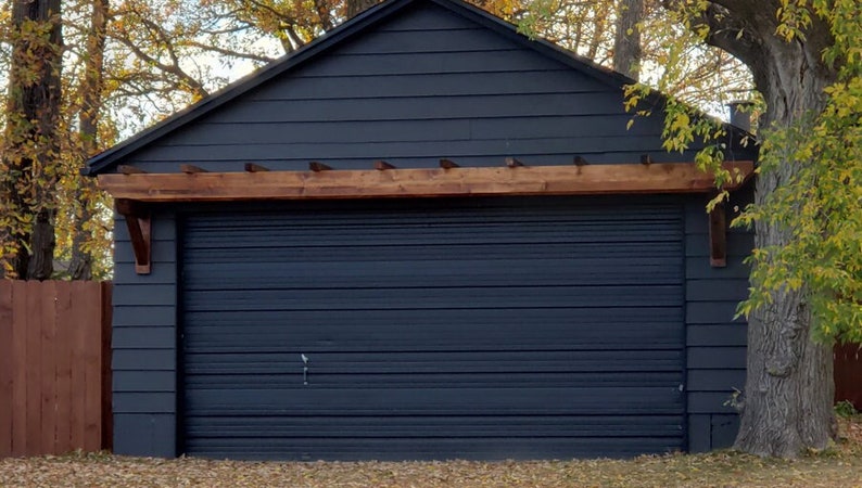 May include: A black garage door with a wooden pergola above it. The garage door is closed and has a wood grain texture. The pergola is made of dark brown wood and has a simple design.