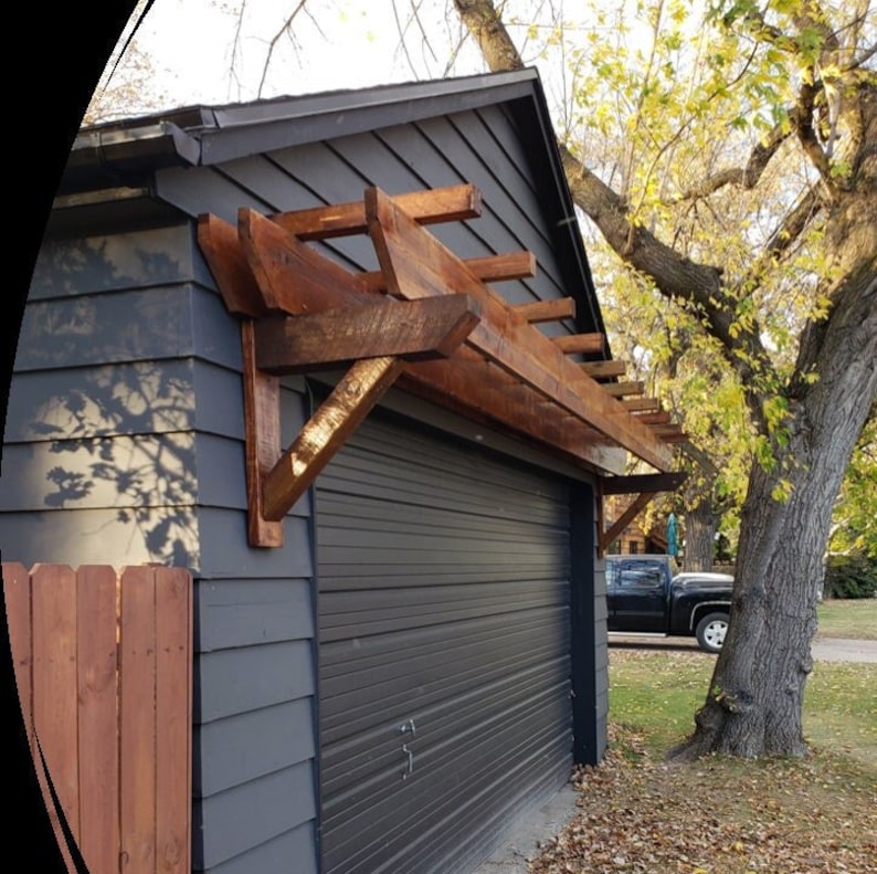 May include: A wooden pergola attached to the side of a gray garage. The pergola is made of natural wood with a brown stain. The garage door is closed and has a gray finish.