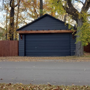 May include: A dark gray garage door with a wooden pergola above it. The garage is surrounded by trees with fall foliage.
