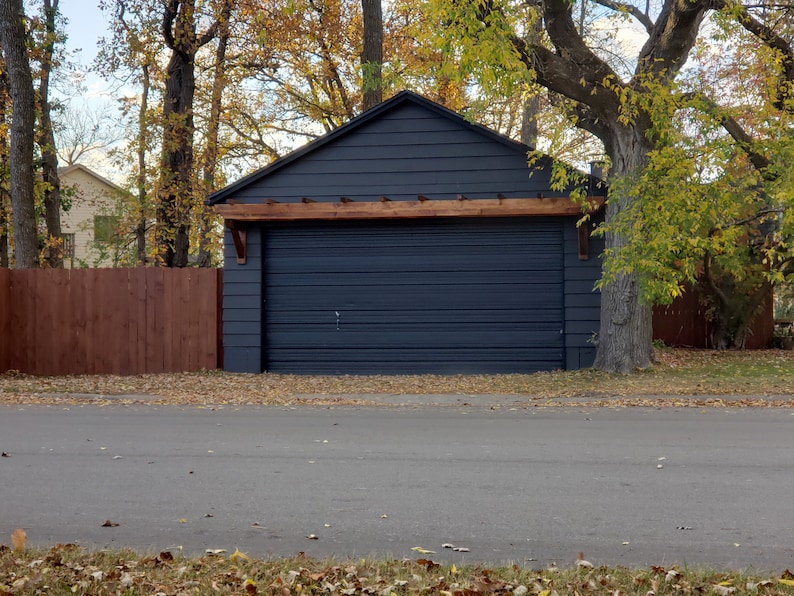 May include: A dark gray garage door with a wooden pergola above it. The garage is surrounded by trees with fall foliage.