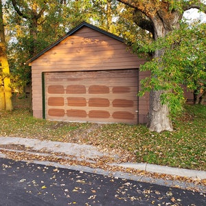May include: A brown wooden garage door with a brick pattern painted on it. The garage is attached to a brown wooden building with a green roof. The garage is surrounded by trees and grass.