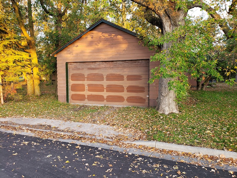 May include: A brown wooden garage door with a brick pattern painted on it. The garage is attached to a brown wooden building with a green roof. The garage is surrounded by trees and grass.