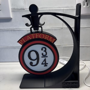 May include: Black 3D-printed sign featuring the words "PLATFORM" and "9 3/4" in a circular design with red and gold accents. The sign is supported by a black stand and has a curved arm. The base of the stand has the text "Harry Potter".