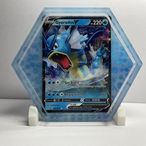 May include: A blue and yellow Gyarados Pokemon card with a holographic finish. The card is encased in a clear hexagonal acrylic display stand. The card features the text "Gyarados V" and "HP 220".