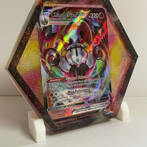 May include: A hexagonal resin display featuring a Chandelure VMAX Pokemon card. The card is set against a colorful background with a black and glittery border. The display is supported by a white stand.
