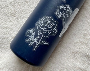 May include: A dark blue water bottle with white floral stickers. The stickers feature a rose, a tulip, and a peony.