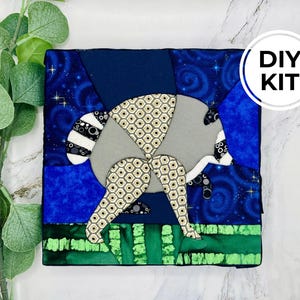 May include: A square fabric quilt block featuring a stylized raccoon appliqué against a dark blue starry night sky and a green grassy foreground. The raccoon is constructed from patterned beige and grey fabric. A white circular label reads DIY KIT.