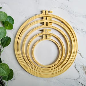 Op de afbeelding: Vier houten borduurringen van verschillende maten zijn gerangschikt op een marmeren oppervlak. De ringen hebben een lichte, natuurlijke houtkleur en elk heeft een metalen spanmechanisme. Een groene bladplant staat op de achtergrond.