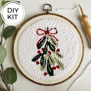 Op de afbeelding: DIY-kit voor een kerstthema punch needle borduurproject. De kit bevat een houten borduurring met witte stof, groen en rood garen en een punch needle tool.