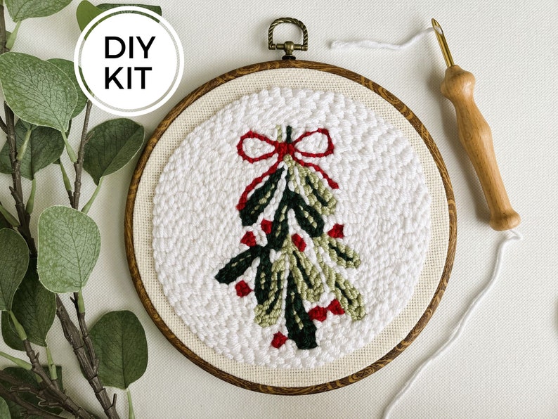 Punch Needle Mistletoe Wall Hanging in Red, Green and white. Shown in faux wood display hoop. Shown next to wood punch needle tool.