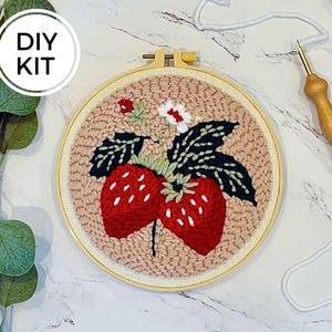 May include: DIY kit for punch needle embroidery featuring a red and white strawberry with green leaves and white flowers on a beige background. The kit includes a wooden hoop, embroidery needle, and yarn.
