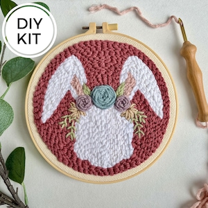 May include: DIY kit for punch needle embroidery featuring a white bunny with pink and green floral accents on a red background. The kit includes a wooden hoop, yarn, and a needle.